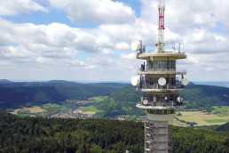 Foto eines Funkmasten mit Bäumen, Wiesen und einigen Häusern im Hintergrund